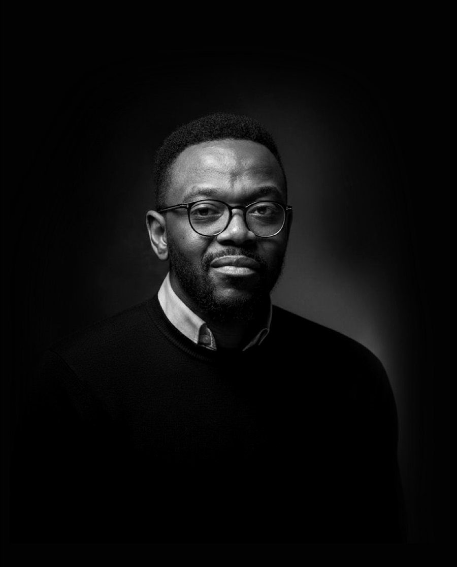 Portrait en noir et blanc de Rémy Muzembo : lunettes, pull sombre sur col clair, fond noir.